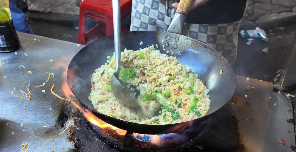Street food vendor stir-frying rice and leafy greens over a high flame