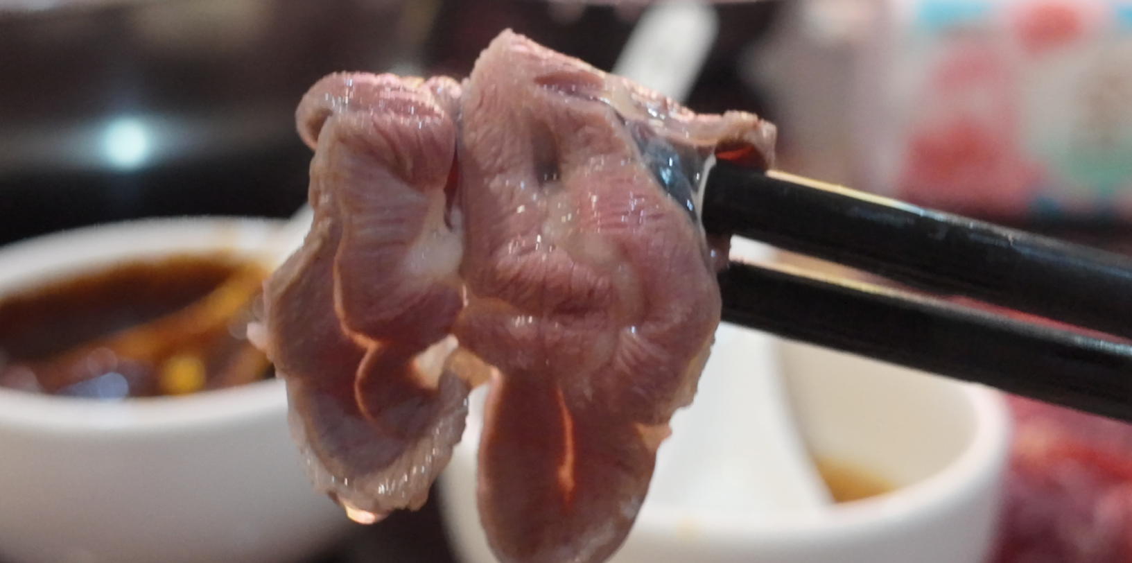 Cooked slice of beef held by chopsticks above a dipping bowl